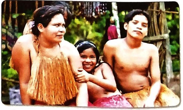A woman in a fringed garment, a smiling girl with a headband, and a shirtless young man are posed outdoors. The background suggests a natural environment with vegetation and wooden structures.