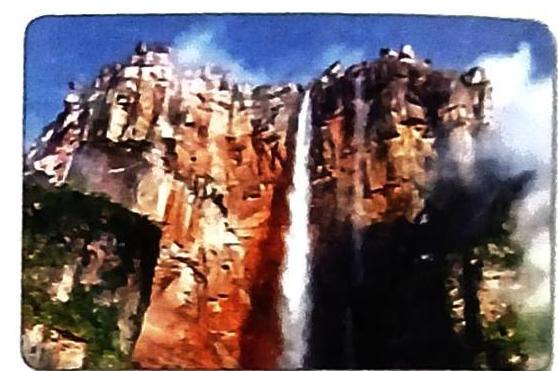 A tall waterfall cascades down a steep, reddish-brown rocky cliff face with patches of green vegetation. Mist surrounds the base of the falls and billows upwards, obscuring parts of the cliff and creating a hazy effect against a blue sky.