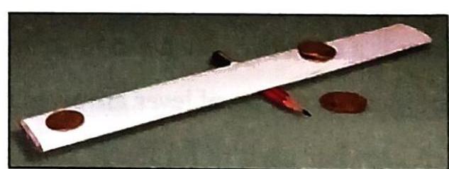 A long, flat, metallic object lies diagonally on a dark surface, with three oval brown objects placed on it at intervals. A red pencil rests beneath the metallic object, with its tip touching it. A fourth oval brown object is located to the right of the metallic object.