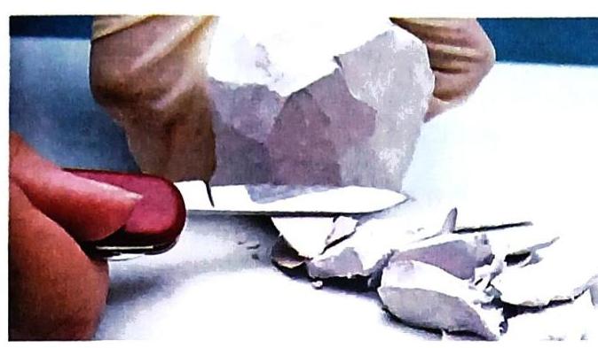 A gloved hand holds a large, faceted crystalline rock while another hand uses a red-handled knife to chip off small pieces.
