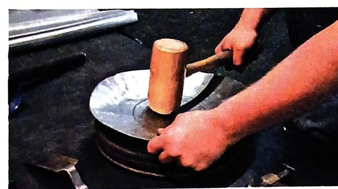 A person's hands hold a wooden mallet above a metal disc resting on a dark surface. Another hand is visible near the disc, and tools lie on the surface.