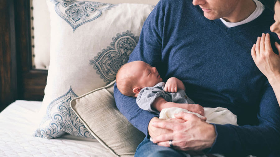 Father holding baby in arms while sitting on couch and mother is smiling