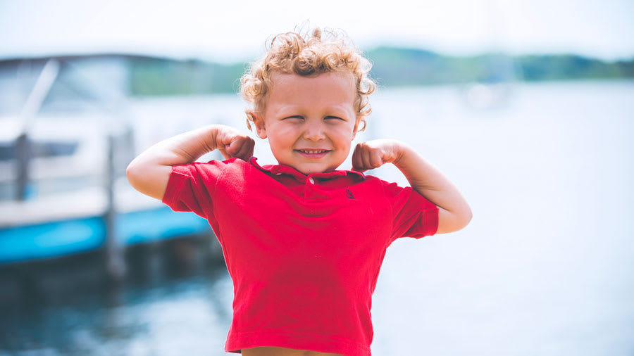 small boy child with short curly hair wearing red polo shirt and blue plaid shorts showing his muscles