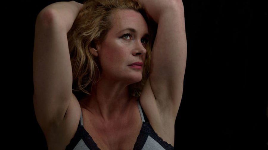 middle-aged blonde woman wearing grey bra with black lace lining holding her arms above her head exposing her armpits and looking sad