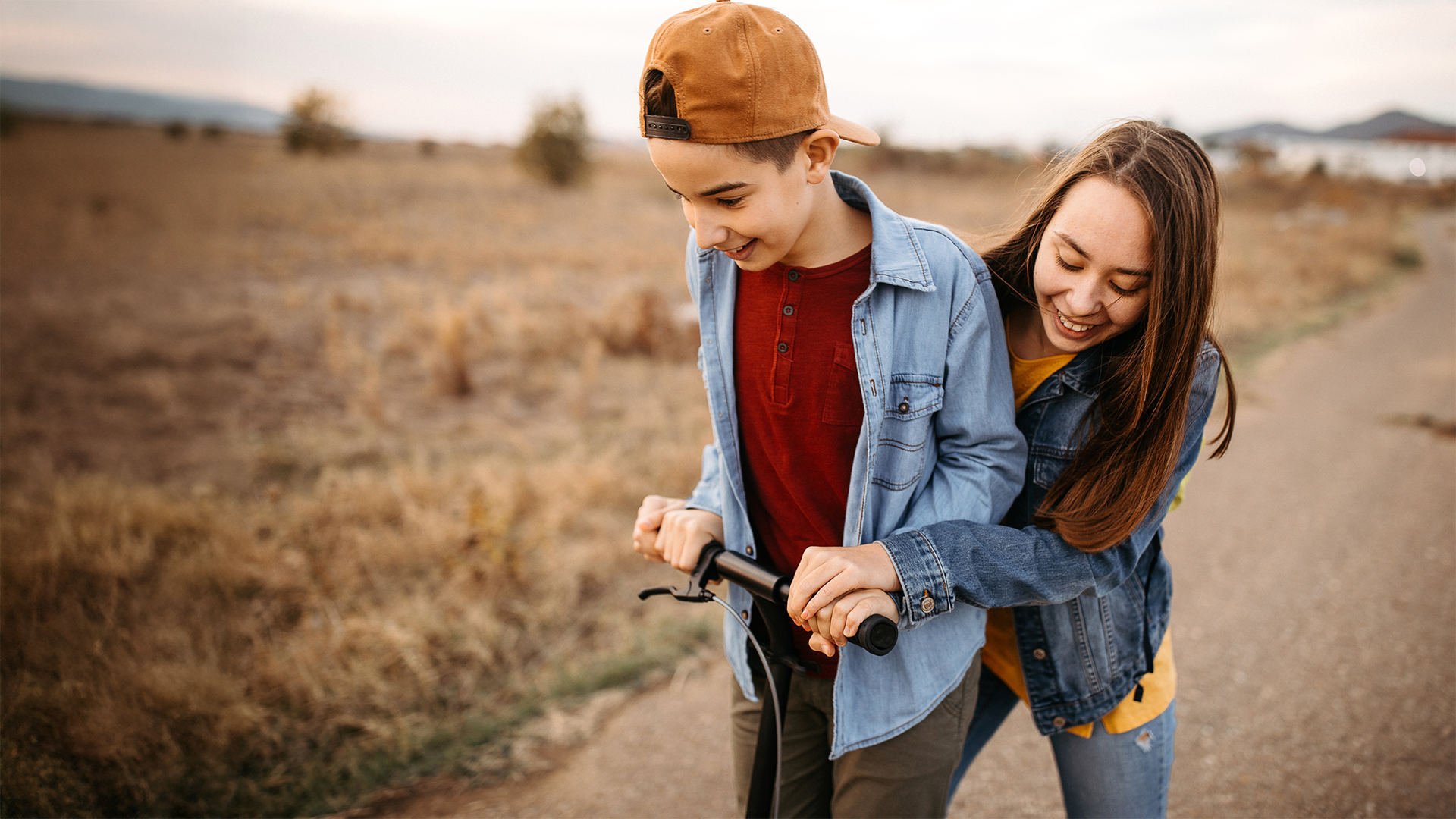 Zwei Jugendliche, Bruder und Schwester, genießen gemeinsam lachend und fröhlich einen Ausflug im Freien. Der Junge fährt auf einem Roller, während das Mädchen ihn unterstützt.