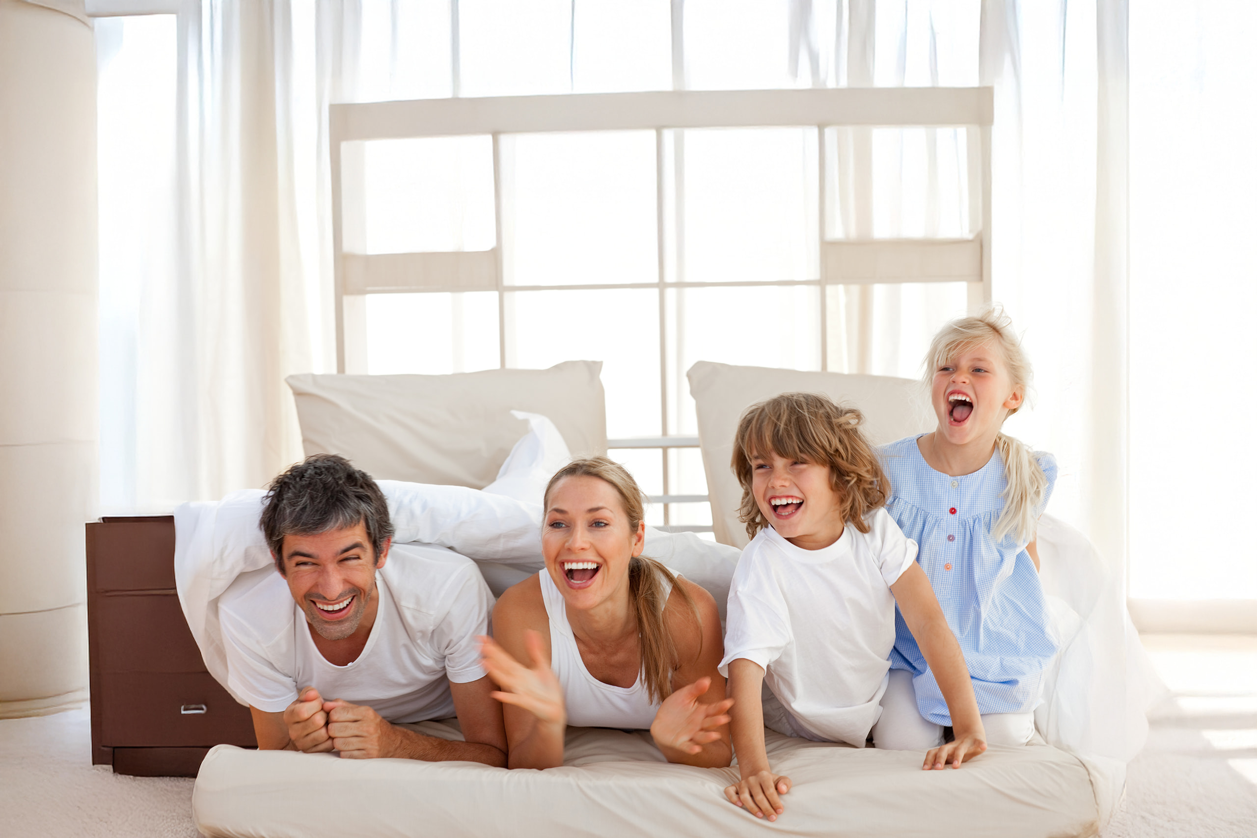 Glückliche vierköpfige Familie mit zwei Kindern, die gemeinsam auf einem Bett lachen und spielerisch Zeit miteinander verbringen. Die Szene wirkt hell, freundlich und vermittelt Lebensfreude und familiäre Verbundenheit.