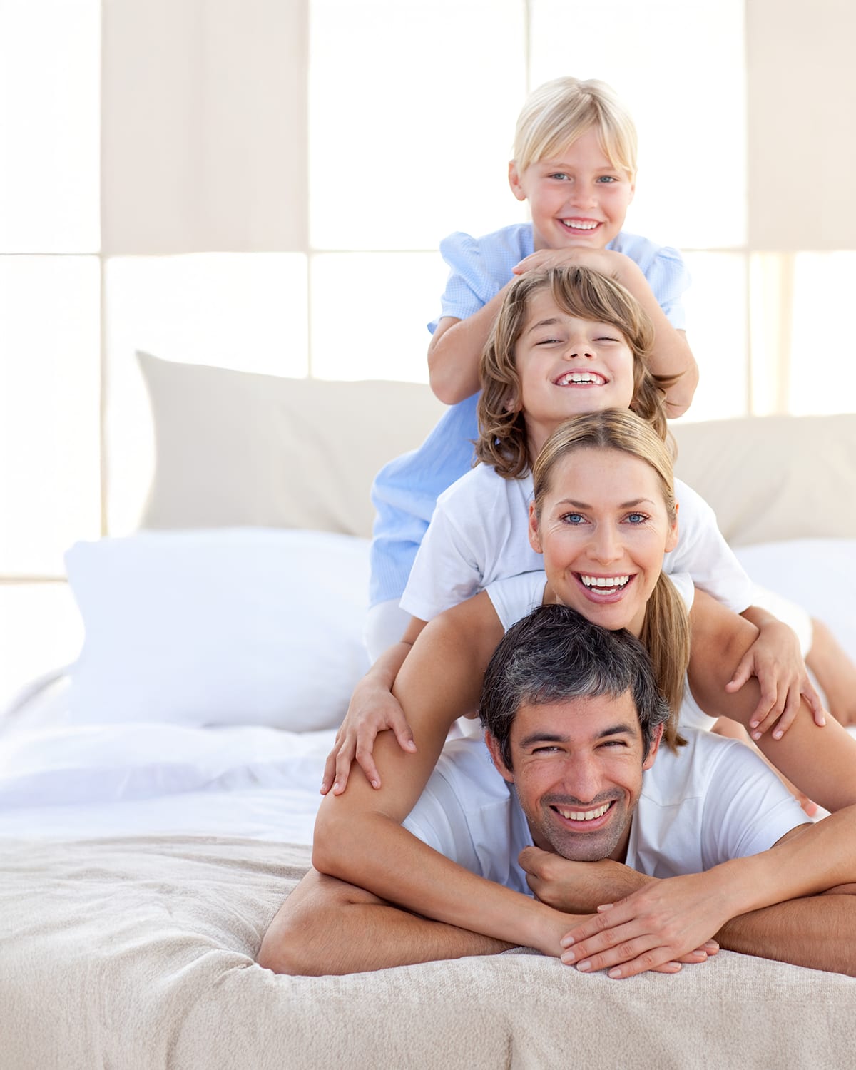 Glückliche vierköpfige Familie mit zwei Kindern, die gemeinsam auf einem Bett lachen und spielerisch Zeit miteinander verbringen. Die Szene wirkt hell, freundlich und vermittelt Lebensfreude und familiäre Verbundenheit.