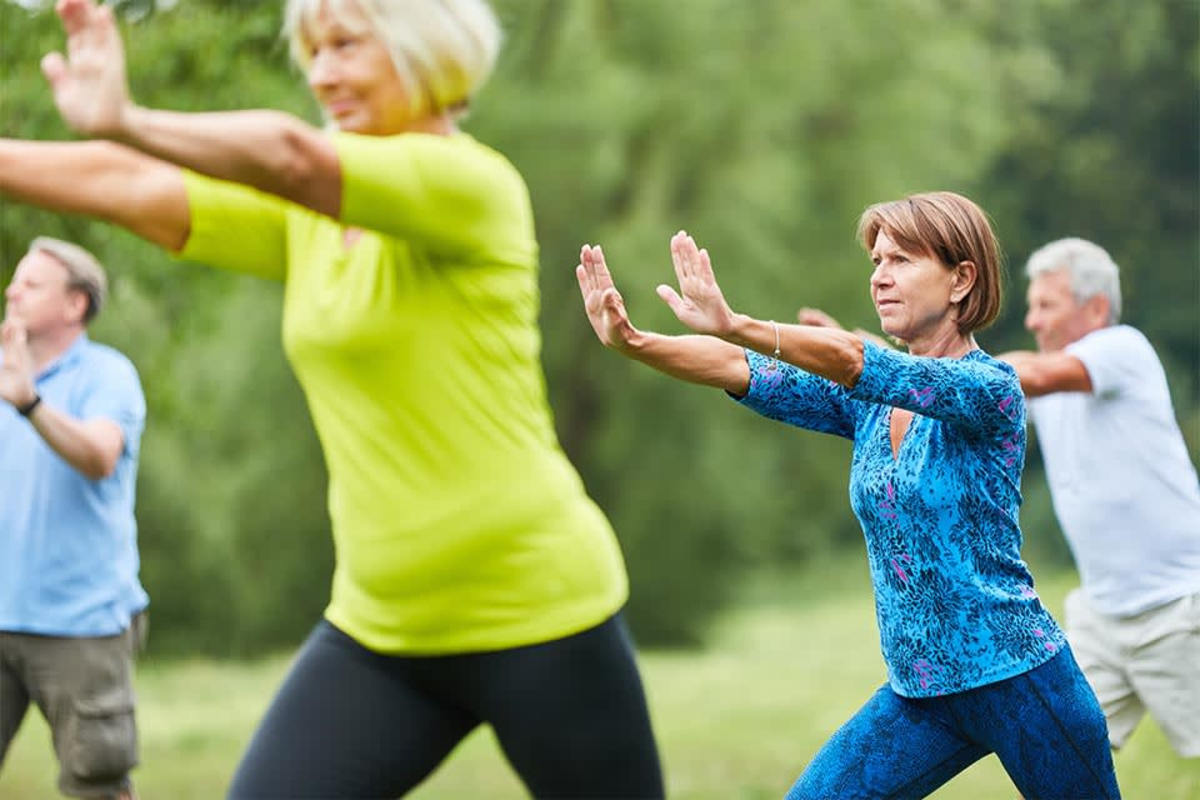 Eine Gruppe von vier Erwachsenen mittleren Alters praktiziert im Freien Tai Chi. Das Bild veranschaulicht Faszientraining in der Natur zur Steigerung von Balance, Körperhaltung und Wohlbefinden.