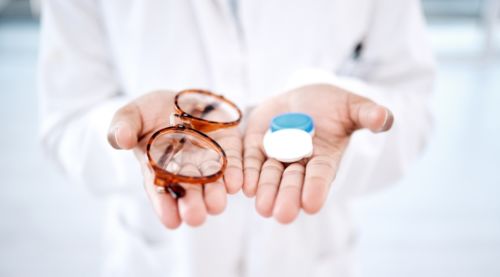 A woman holding a pair of glasses in one hand and contact lenses in the other, showcasing both options for vision correction.