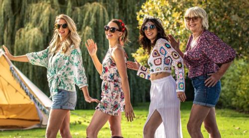 Four women dancing outside in summer clothing and sunglasses, enjoying a joyful moment together under the sun.