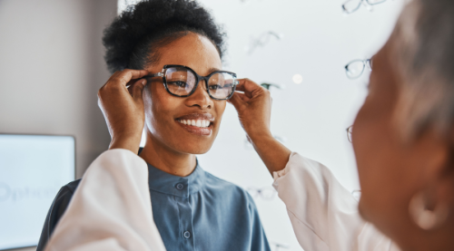 Optician conducting a glasses fit check appointment with a woman patient, adjusting lenses while placing glasses on her face.