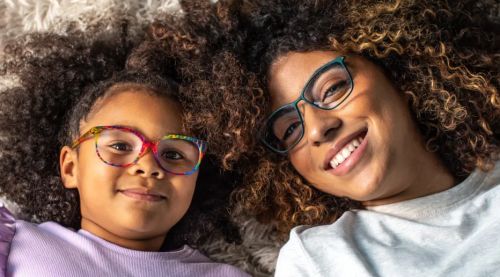 A mother and her daughter smiling while lying on the floor, both wearing stylish glasses and enjoying a playful moment together.