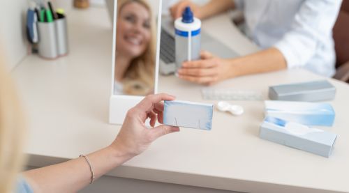 Close-up of a female hand holding a small blue and white box containing contact lenses, with a doctor holding a bottle of lens solution in the blurred background.