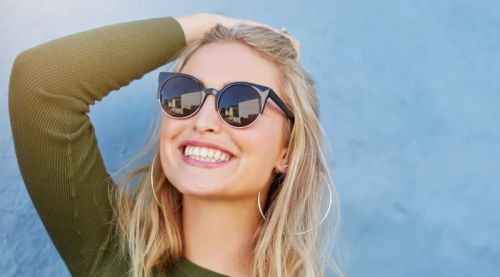 A smiling woman wearing glasses, resting her hand on her hair, exuding confidence and joy.