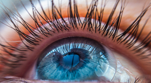 Extreme close-up of a blue human eye wearing a contact lens, looking upward, showcasing the intricate details of the iris and lens.
