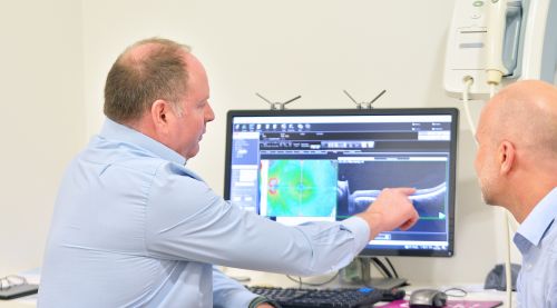 An eye care clinician explaining OCT (Optical Coherence Tomography) scan images to a patient, pointing out specific retinal layers on a computer screen.