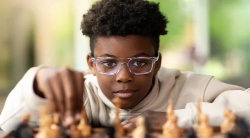 A smiling child wearing glasses, focused on playing chess, with pieces arranged on the board in front of them.