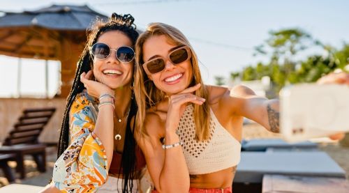 Smiling women on beach in summer, wearing sunglasses, using smartphone taking selfie picture.