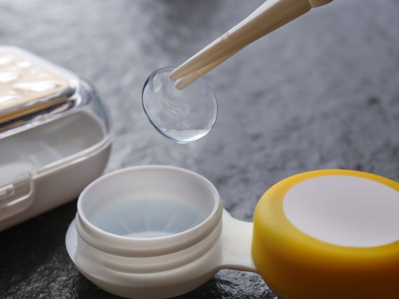 Tweezers holding a contact lens, with a plastic container, on a gray background.