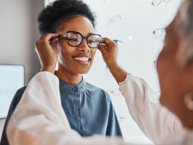 Optician conducting a glasses fit check appointment with a woman patient, adjusting lenses while placing glasses on her face.