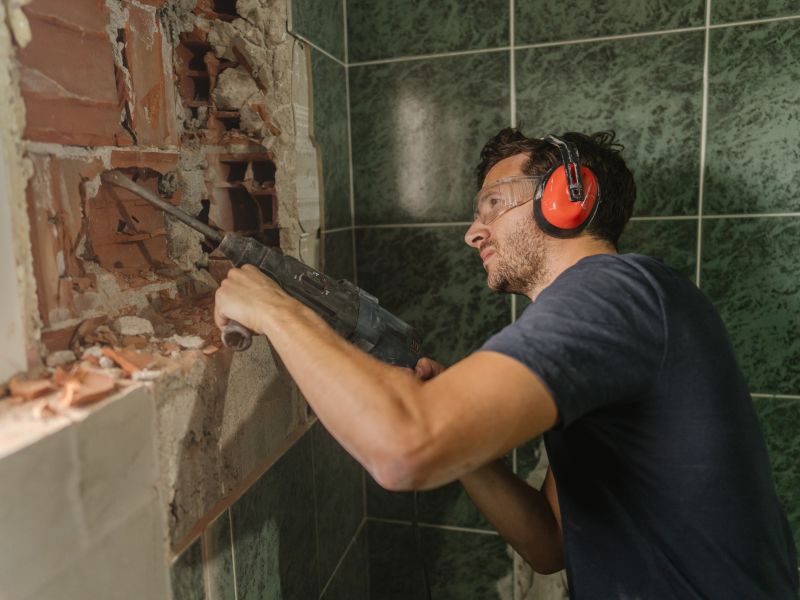 A man wearing hearing protection and eye protection using a jackhammer to break a wall.