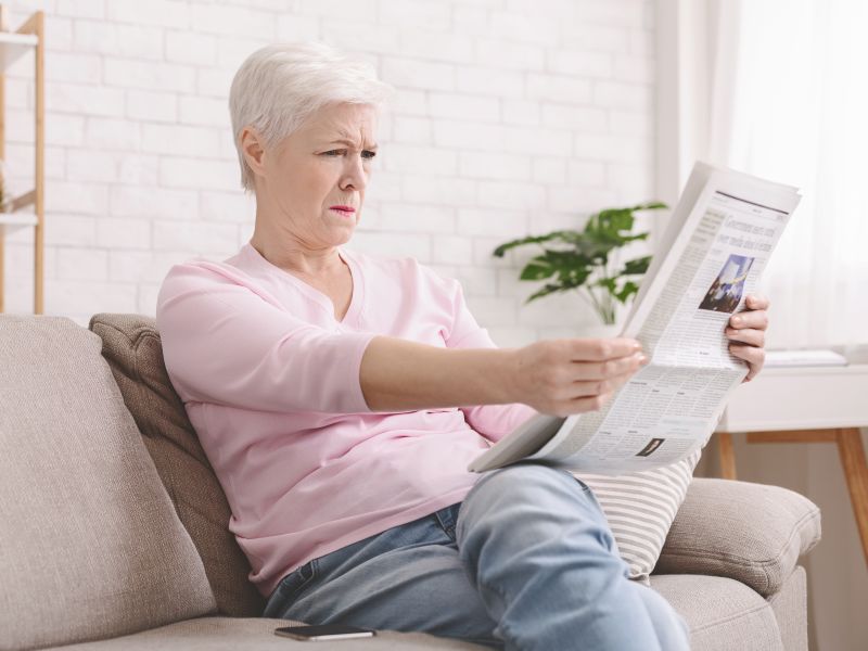 A senior woman holding a newspaper at arm's length and squinting.