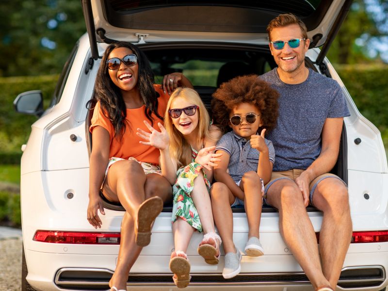 A happy family wearing sunglasses, sitting on their car boot, and smiling.