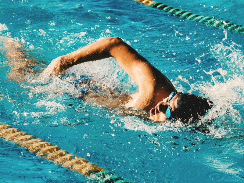 Man swimming wearing a hat and goggles in an indoor public swimming pool.