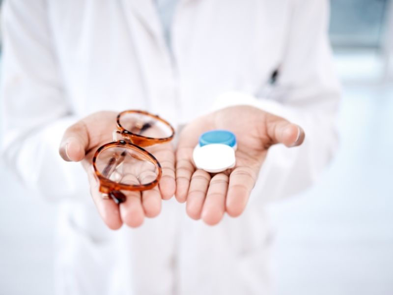 A woman holding a pair of glasses in one hand and contact lenses in the other, showcasing both options for vision correction.