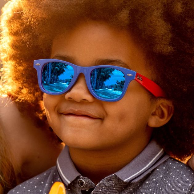 A young boy wearing Ray-Ban Junior, holding a glass of orange juice and looking happy.