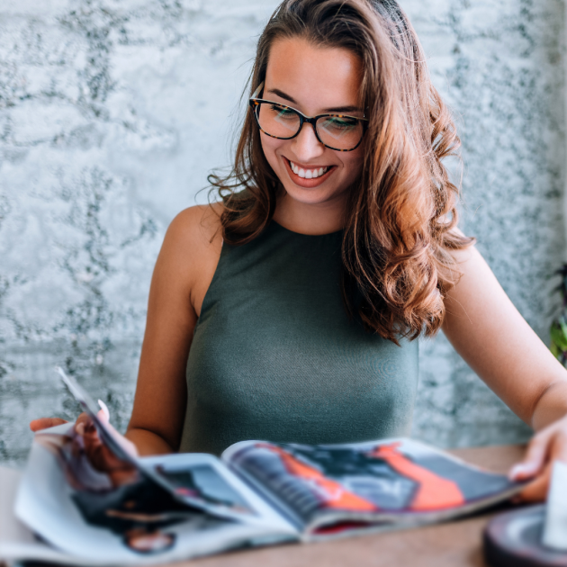 Young girl with glasses reading magazine close to window.