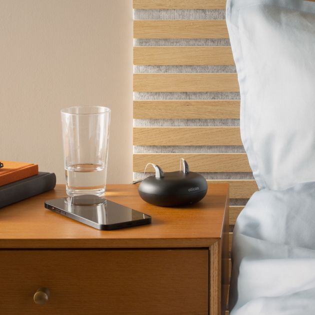 A bedside table with Oticon Real hearing aids placed in the charger.