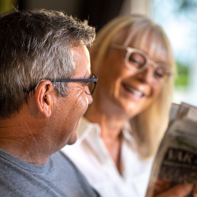 A man wearing hearing aids with a woman wearing glasses in the blurred background.