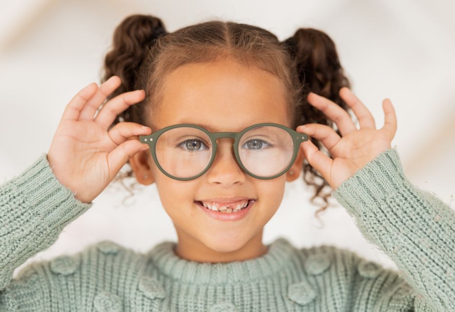 A smiling little girl putting on a pair of green glasses, radiating joy and excitement.