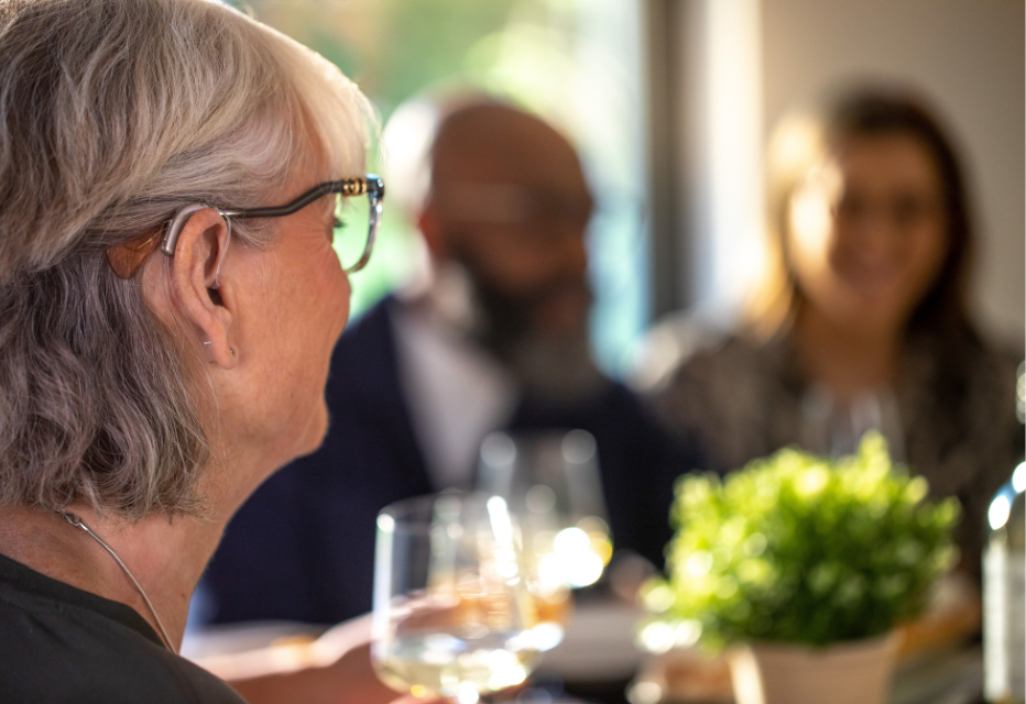 Close-up of a woman wearing a hearing aid in the foreground, while two people in the background are engaged in conversation.