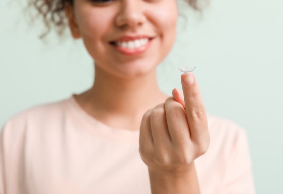 A close-up of a woman's hand holding a contact lens on her index finger, with her smiling face softly blurred in the background.