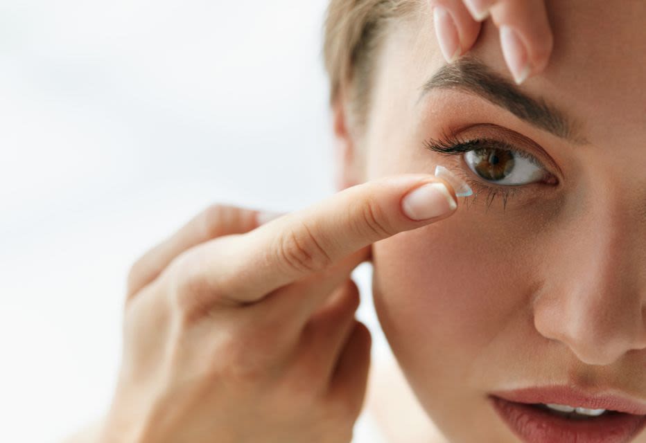Close-up of a woman applying a contact lens to her brown eye, highlighting her focused expression as she gently uses her hands to insert the lens.