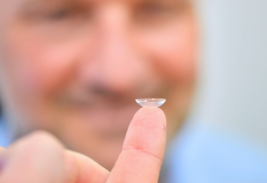 A close-up of a contact lens balanced on a fingertip, with a blurred man's face in the background.