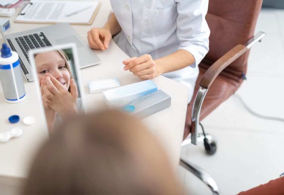 Close-up of a young child carefully inserting a contact lens while holding up her eyelid, with an optician in a white lab coat sitting nearby at a table with a laptop and lens solution bottle.