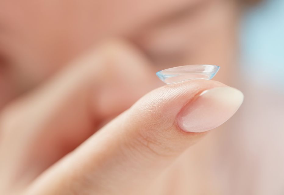 Close-up of a soft contact lens balanced on a woman's fingertip, with her blurred face in the background.