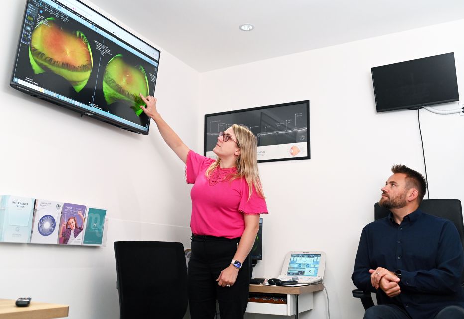 A patient seated in a consulting room while an optometrist stands nearby, explaining the results of an Optical Coherence Tomography (OCT) scan using a screen.