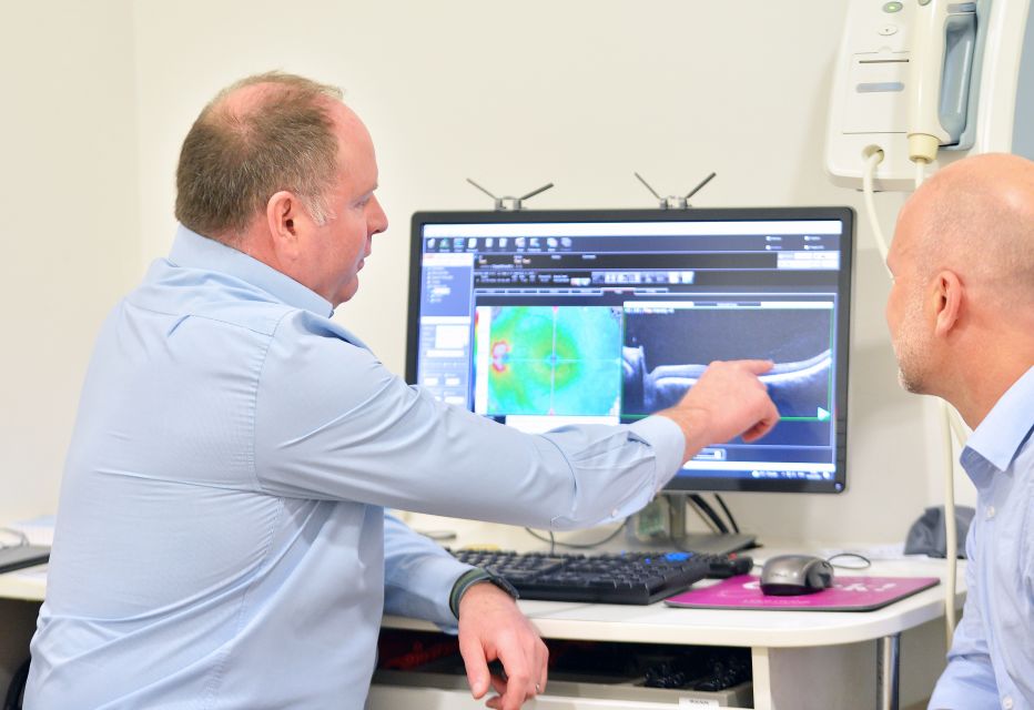 An eye care clinician explaining OCT (Optical Coherence Tomography) scan images to a patient, pointing out specific retinal layers on a computer screen.