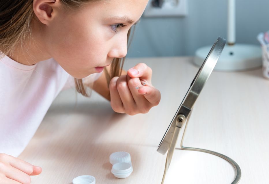 A child applying contact lenses while standing in front of a mirror, focusing on her reflection as she carefully handles the lenses.
