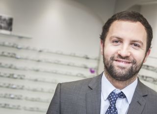 A smiling man with a blurred background of shelves filled with various eyewear.