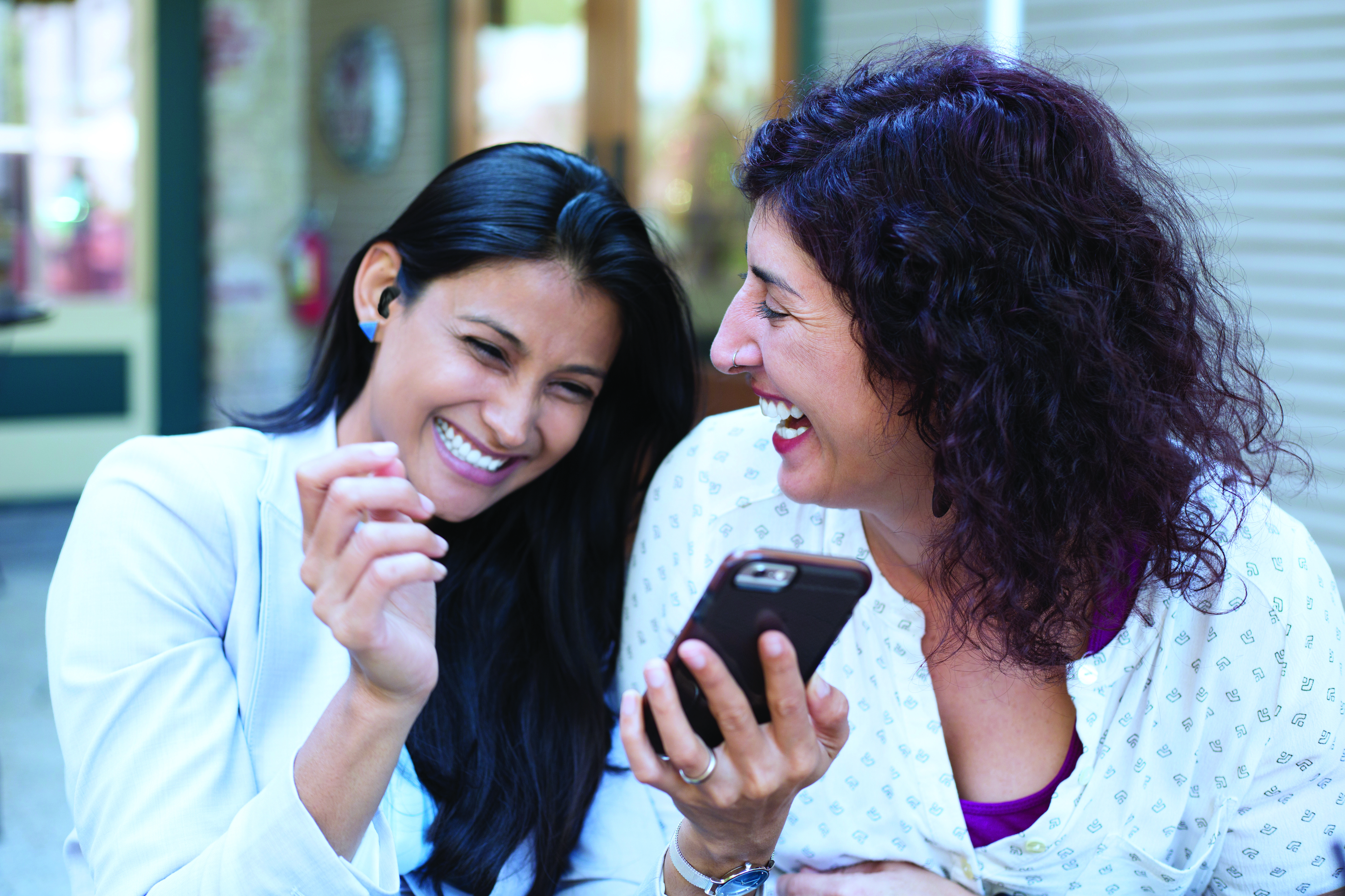 A woman wearing a hearing aid laughs with her friend while looking at her phone.