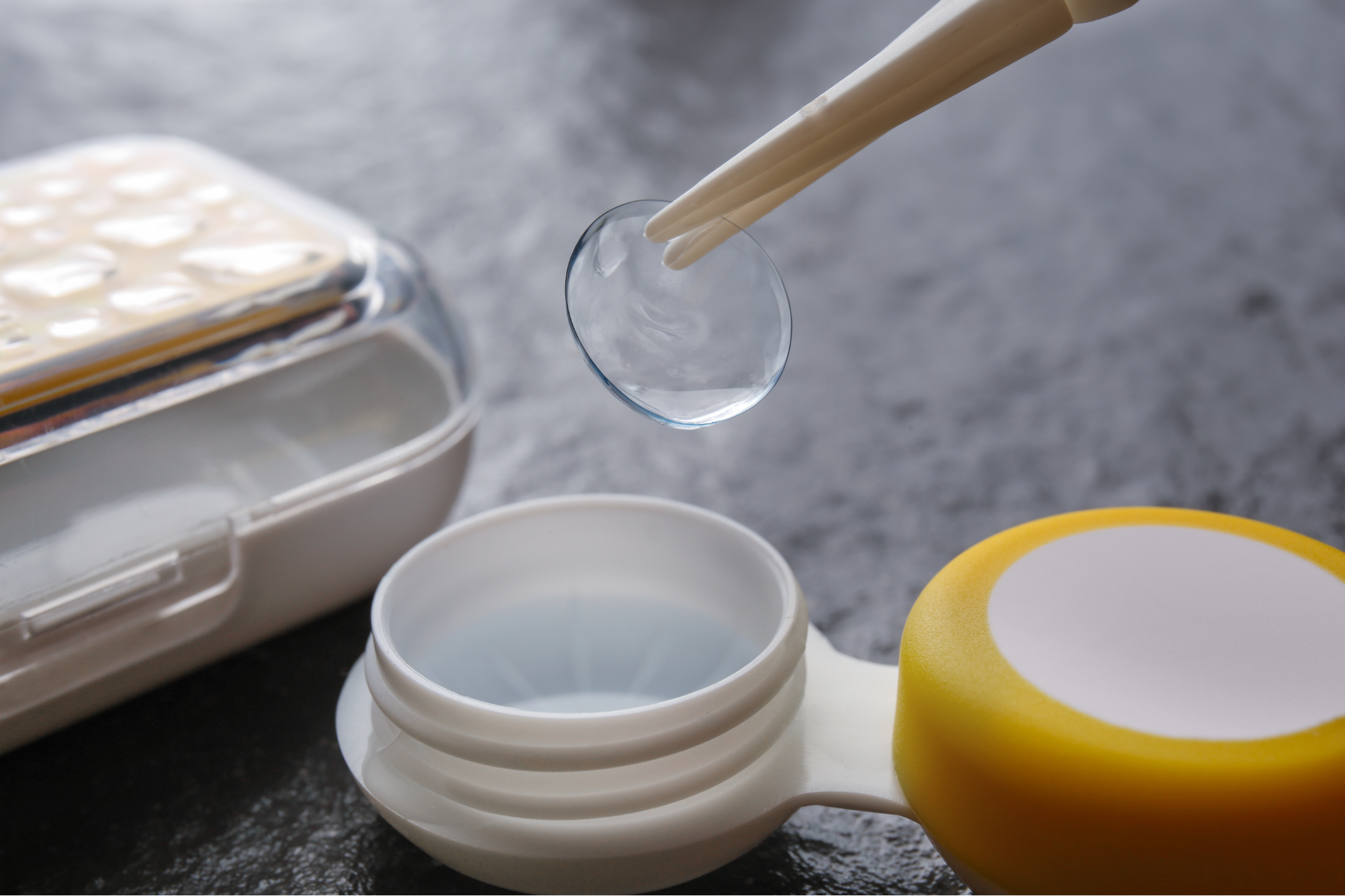 Tweezers holding a contact lens above a plastic container on a gray background, illustrating the careful handling of lenses.