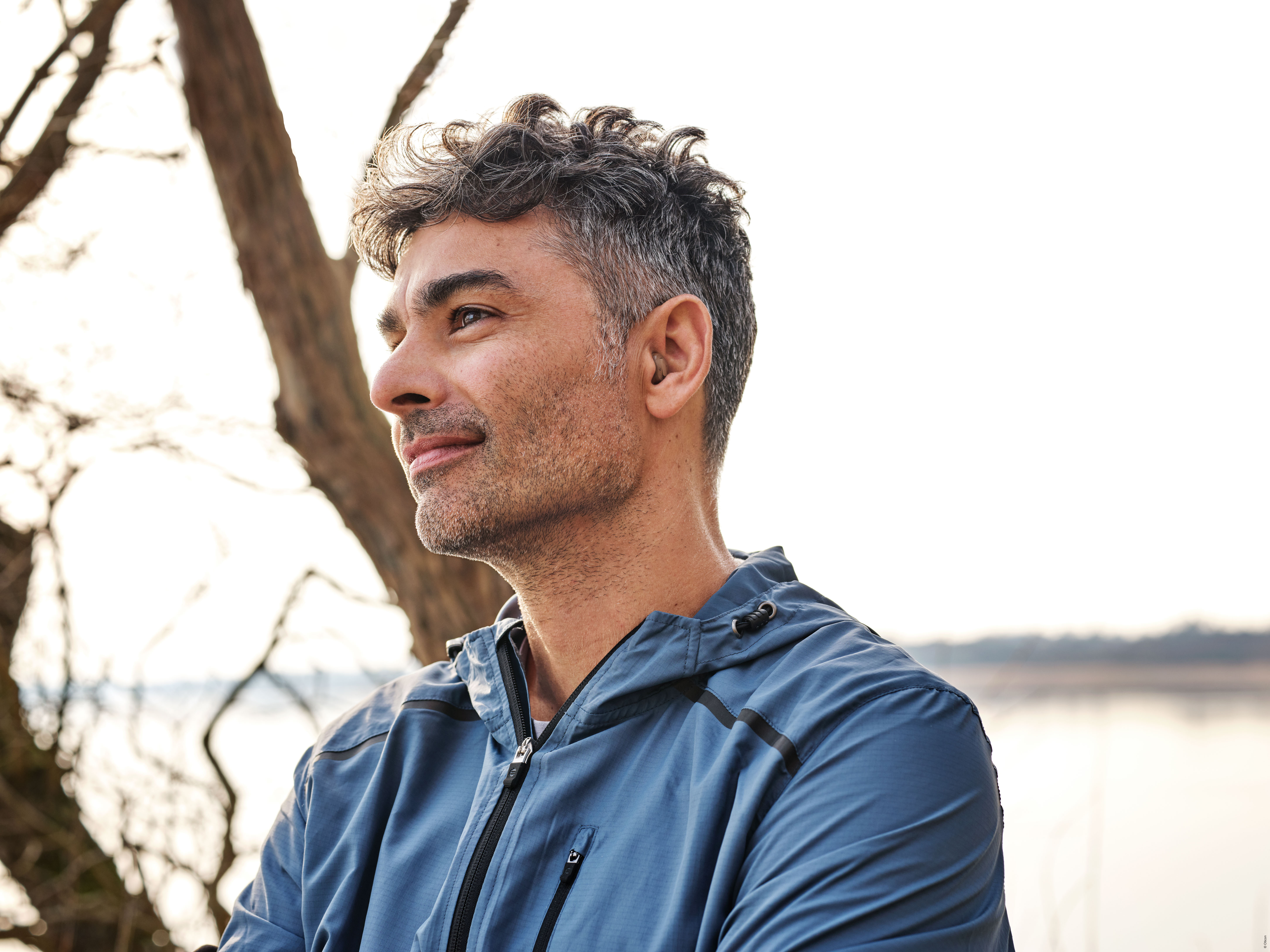 A smiling man wearing an Oticon Own hearing aid in his ear, dressed in sports clothes and enjoying the outdoors.