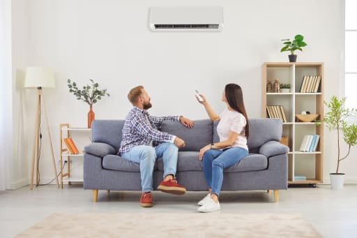 Junges Familienpaar sitzt im Wohnzimmer auf dem Sofa und schaltet die Klimaanlage ein