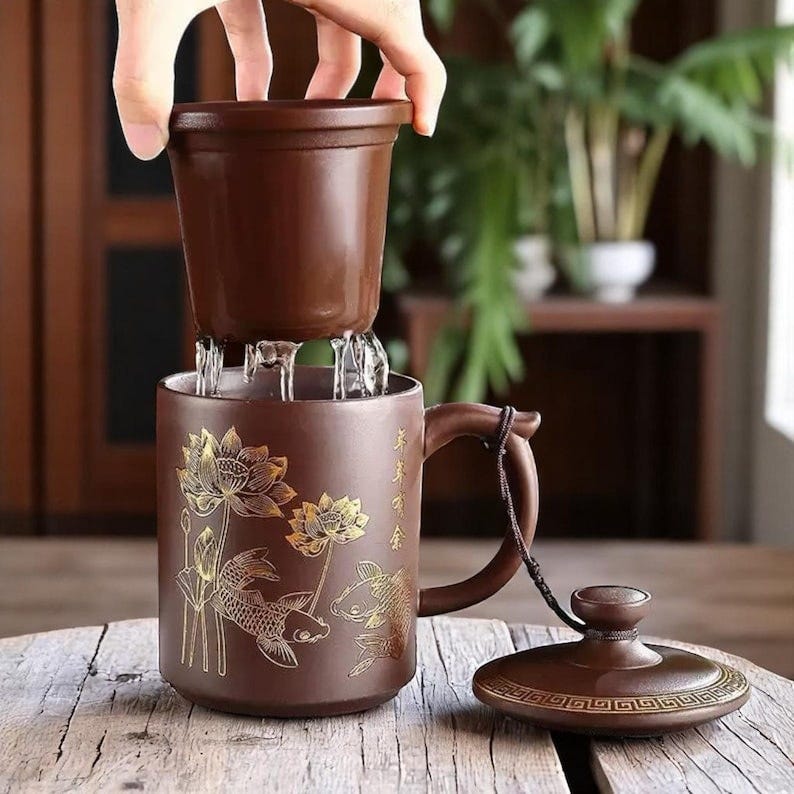 May include: A brown ceramic teacup with a gold lotus flower and fish design. The teacup has a lid and a built-in strainer. The teacup is sitting on a wooden table.