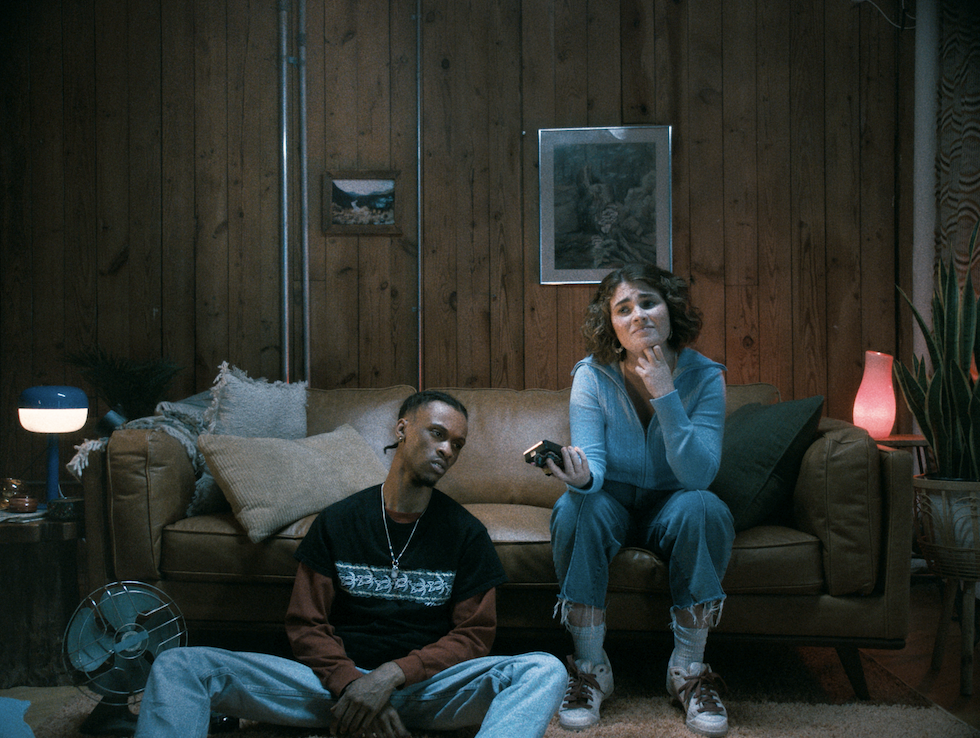 A girl sitting on a sofa, holding her phone, while a guy sits on the ground beside her. Both are listening to something on the phone, focused and engaged.